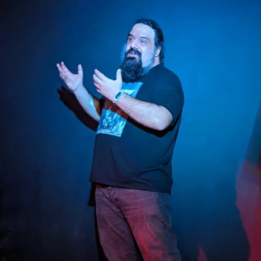Orestis Vasilakis on stage, mid-sentence, lit in deep blue